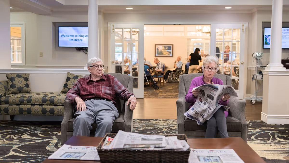 Two older adults sit in armchairs in a senior living residence lounge, one reading a newspaper with other residents visible in a dining area behind them.