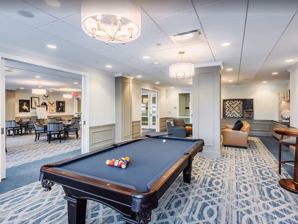Bright communal recreation room with a pool table in the foreground, seating areas, and dining tables in the background.