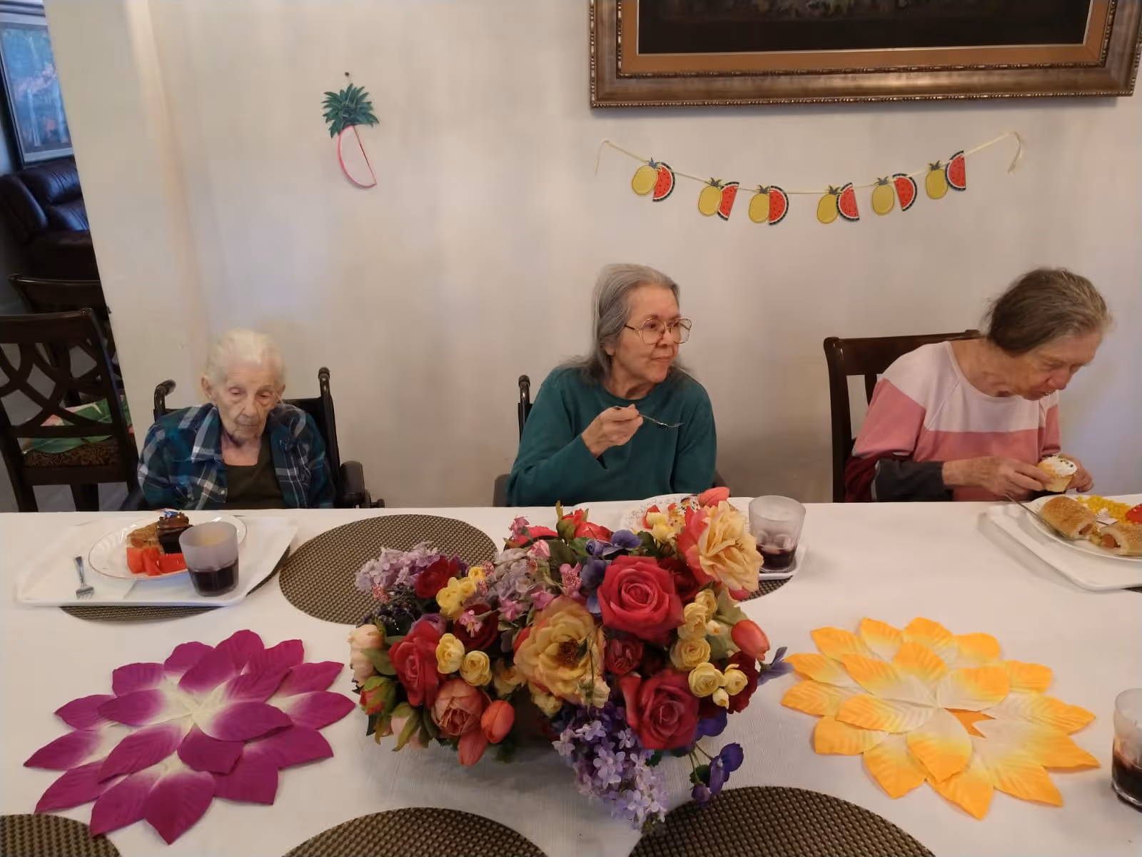 Three elderly women sitting at a dining table in a senior living facility, eating food from trays. The table is decorated with a large colorful flower arrangement and two flower-shaped placemats. The wall behind them has a small palm tree decoration and a fruit-themed garland.