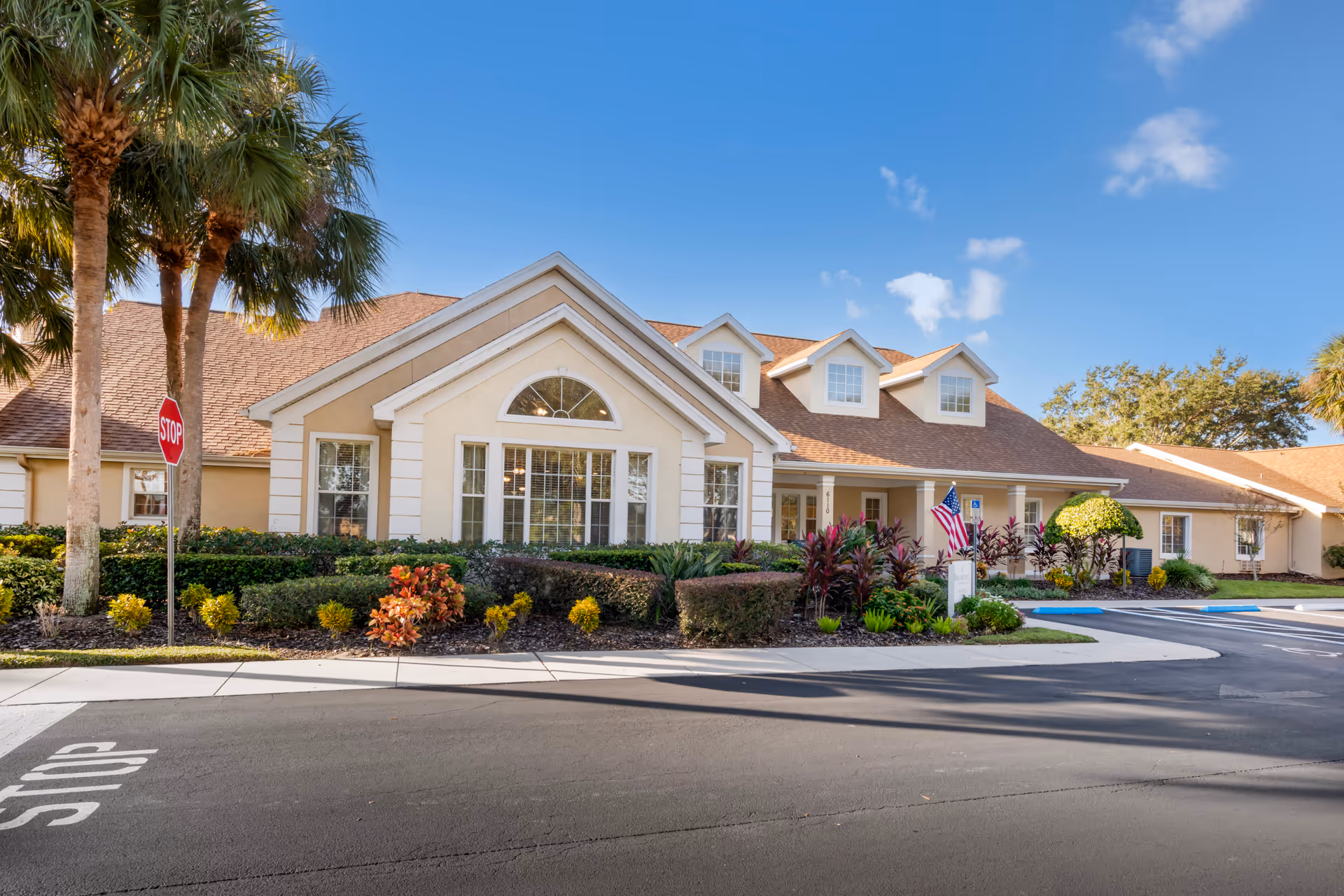 Front exterior of a beige, single-story senior living building with manicured landscaping, palm trees, an American flag, and a stop sign.