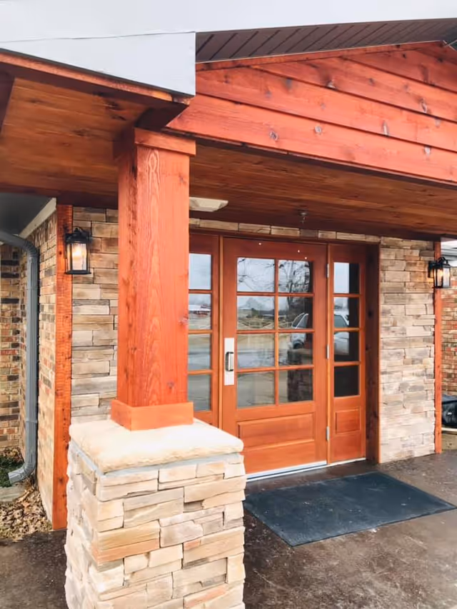 Entrance of a building with wooden double doors featuring glass panes, stone pillars, and a wooden overhang. There are two wall-mounted lantern-style lights on either side of the doors and a black mat on the ground in front of the entrance.