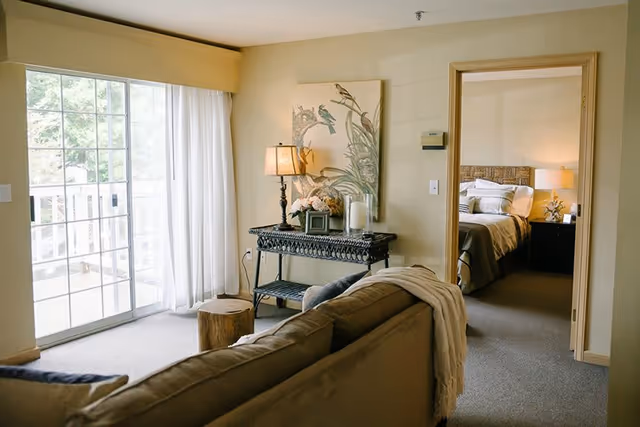 A cozy living room area with a brown sofa draped with a light-colored blanket, a small wooden stump side table, and a decorative console table with a lamp, flowers, and candles. A large window with sheer white curtains lets in natural light. Through an open doorway, a bedroom with a neatly made bed and bedside lamps is visible.