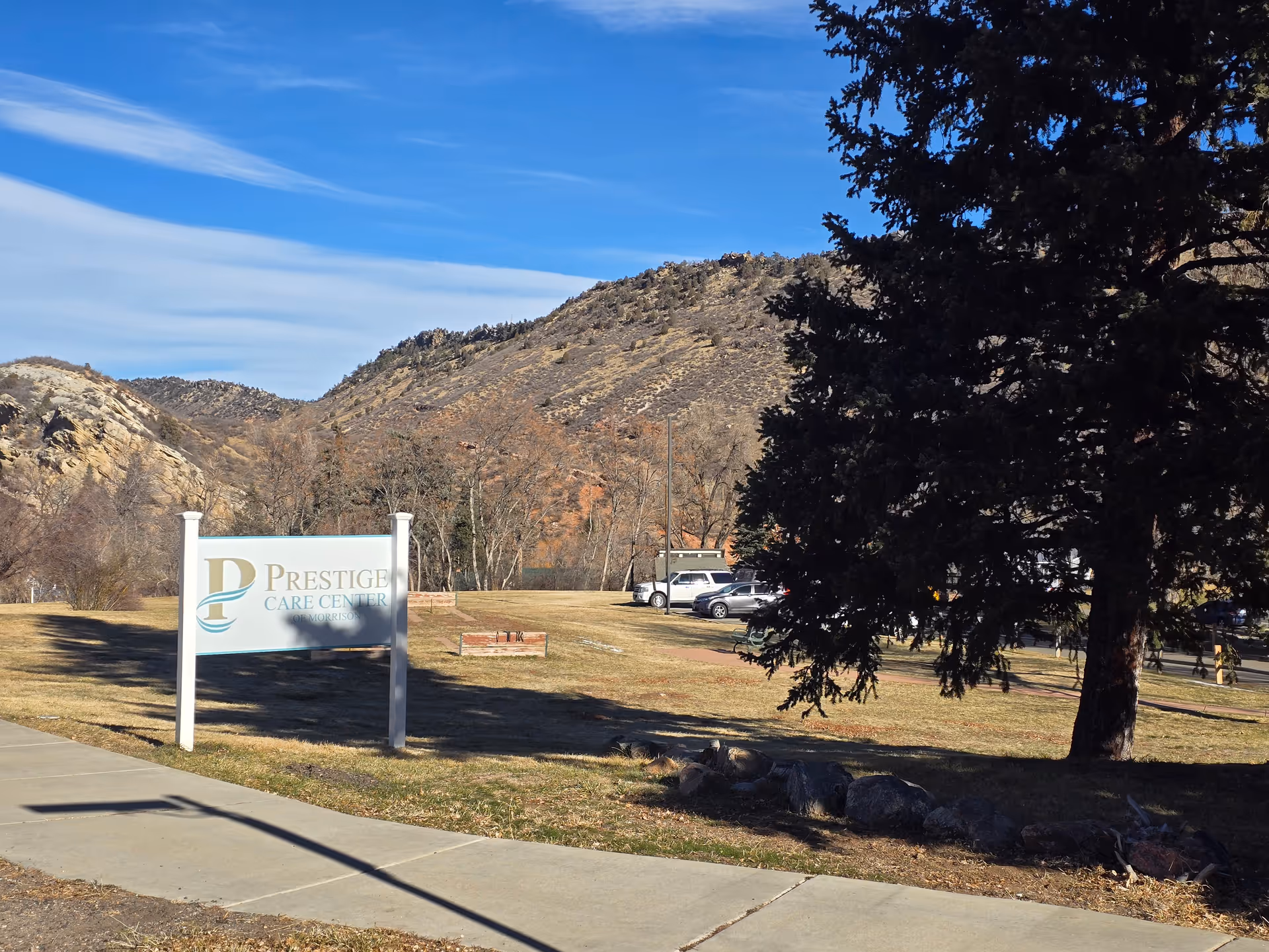 Sign for Prestige Care Center on a grassy lawn with a large tree, parked cars, and rocky hills in the background.