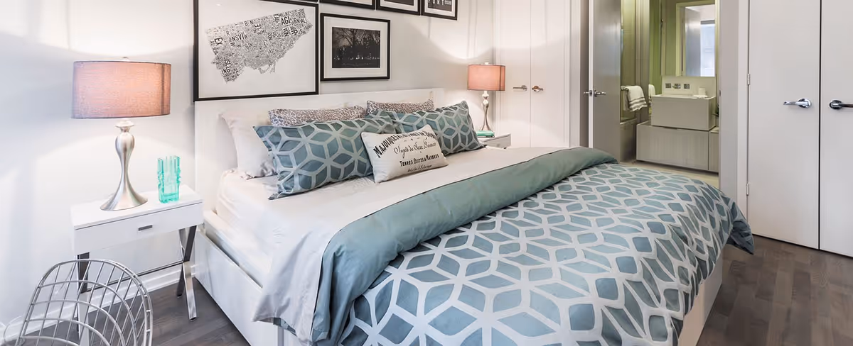 A modern bedroom with a large bed featuring a geometric patterned blue and gray comforter and multiple pillows. There are two bedside tables with lamps on each side of the bed. The wall behind the bed has framed artwork. To the right, there is an open door leading to a bathroom with a sink and mirror visible.