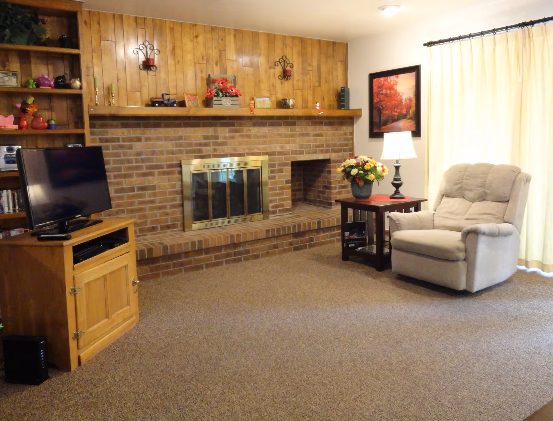 A cozy living room with a brick fireplace and wooden paneling above it. To the left, there is a wooden TV stand with a flat-screen television. On the right side, there is a beige recliner chair next to a wooden side table with a lamp and a vase of flowers. A framed picture of autumn trees hangs on the wall above the side table, and light-colored curtains cover the window.