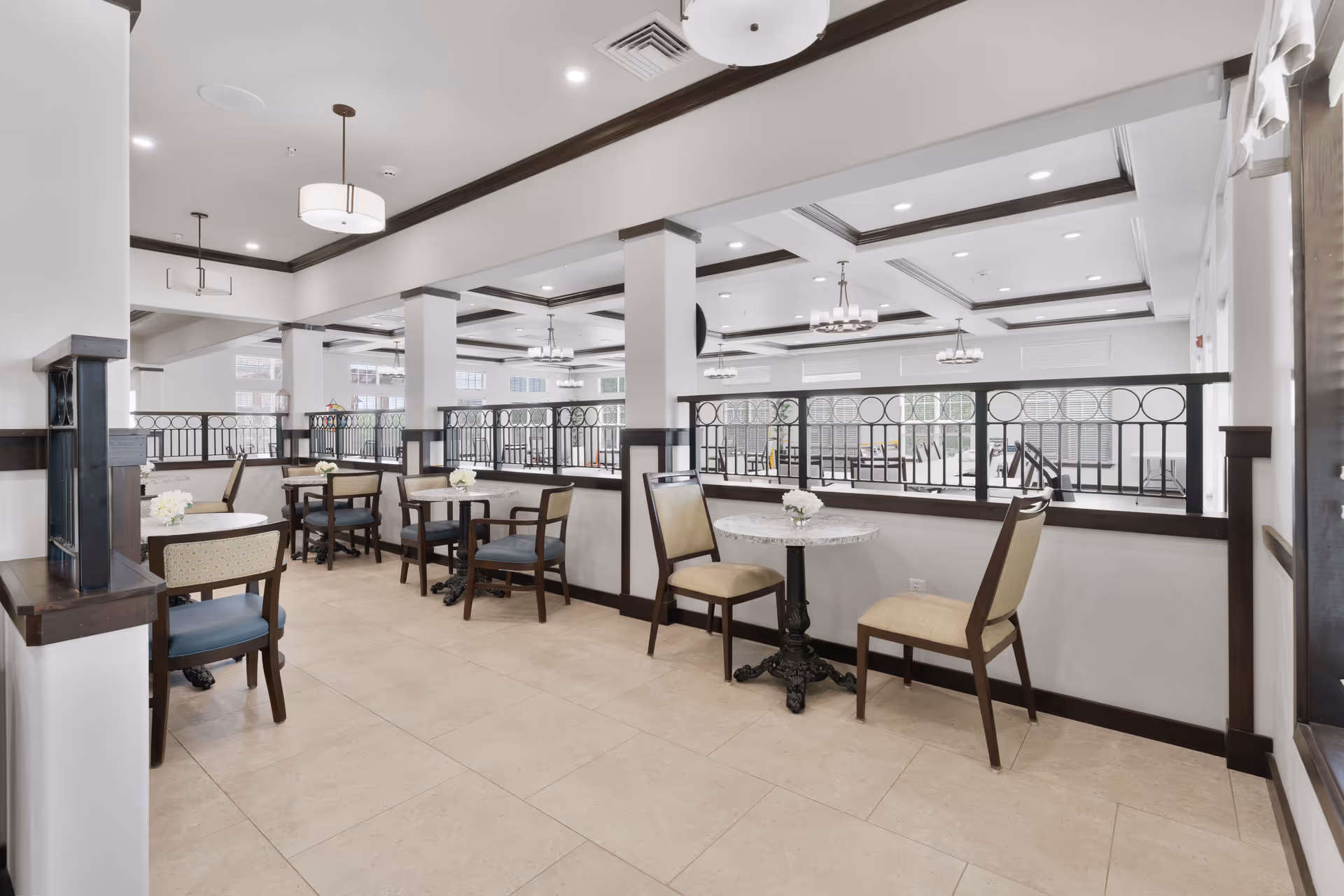A bright and spacious dining area with several small round tables, each accompanied by two chairs. The room features beige tiled floors, white walls with dark wood trim, and large windows allowing natural light to fill the space. Decorative ceiling lights hang from a coffered ceiling, and a black metal railing separates this dining area from another section of the room.