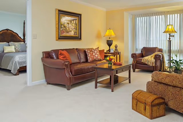 A cozy living room with a brown leather sofa and armchair, a wooden coffee table with a flower vase, a side table with a lamp, and a large window with sheer curtains. To the left, an open doorway reveals a bedroom with a wooden bed frame and neatly made bed.