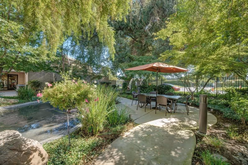 Outdoor garden area at Campus Commons Senior Living with a curved concrete pathway, a table with six chairs under an orange umbrella, surrounded by lush green trees and plants, and a small water feature on the left side.