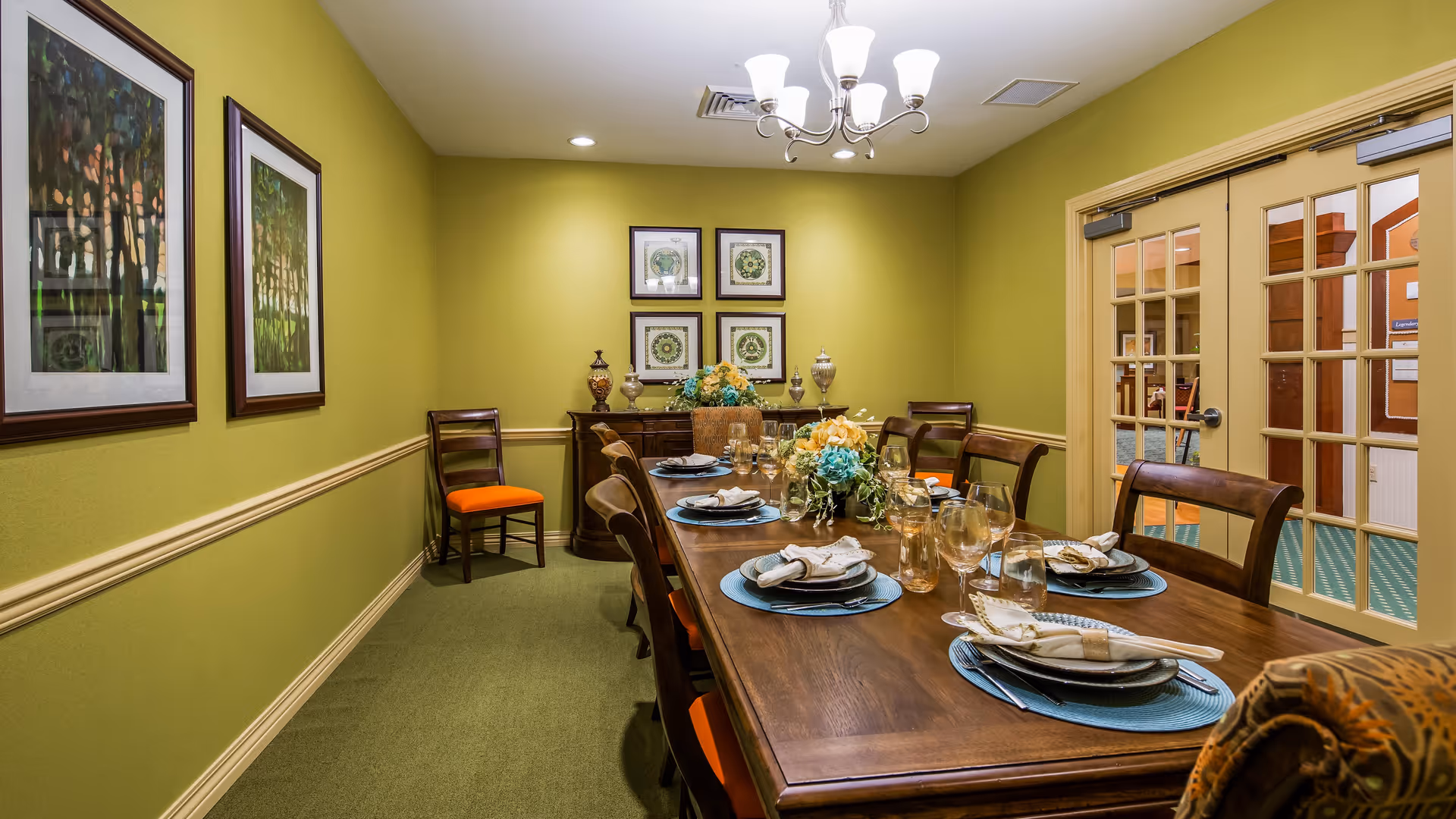 A dining room with a long wooden table set for a meal, featuring plates, napkins, glasses, and floral centerpieces. The room has green walls with framed artwork, a chandelier overhead, and wooden chairs with orange cushions. French doors lead to another room.