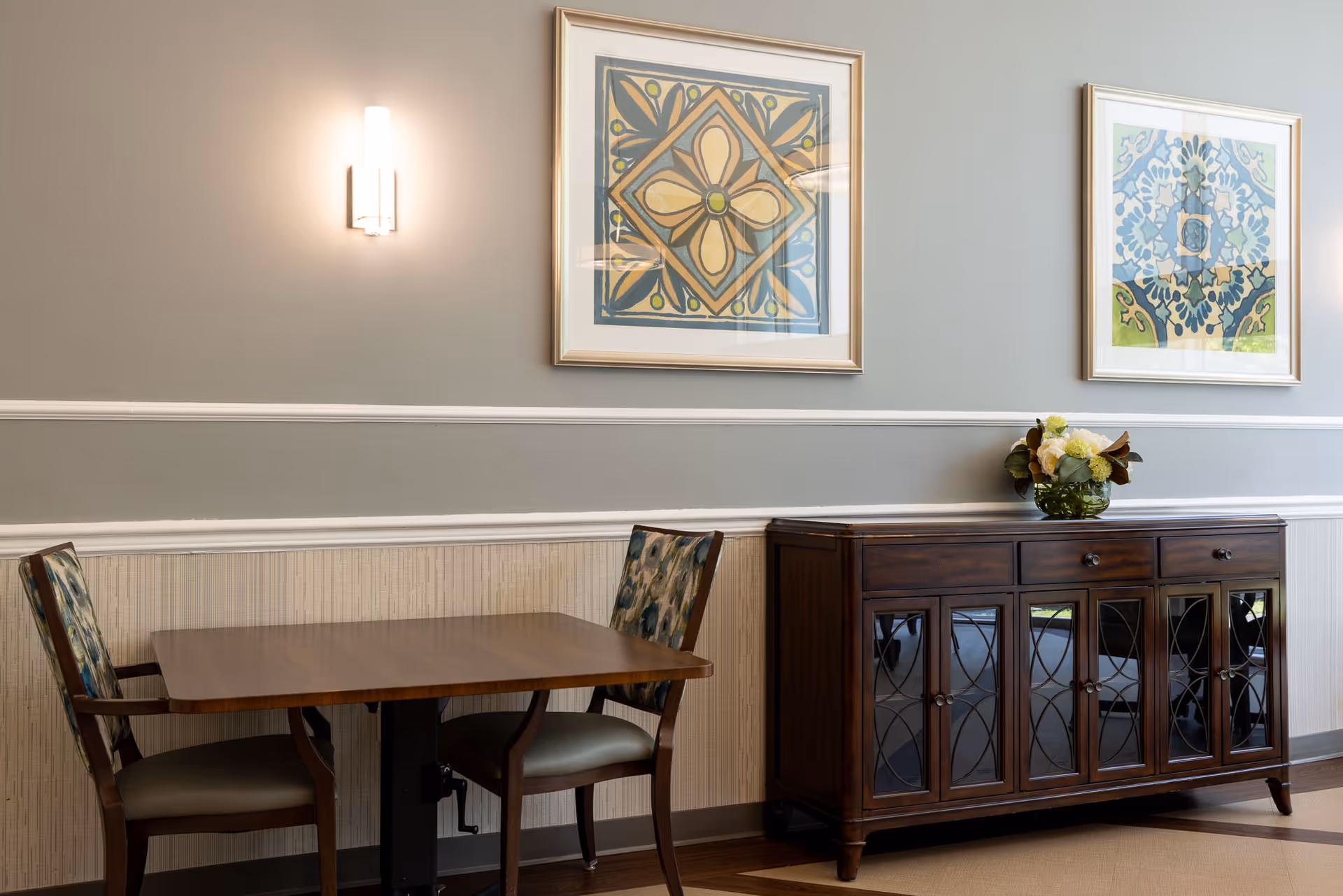 A small dining area with a wooden table and two chairs with patterned upholstery. On the right side, there is a wooden cabinet with glass doors and a flower arrangement on top. Two framed abstract artworks hang on the gray wall above the cabinet and table. A wall-mounted light fixture is also visible above the table.