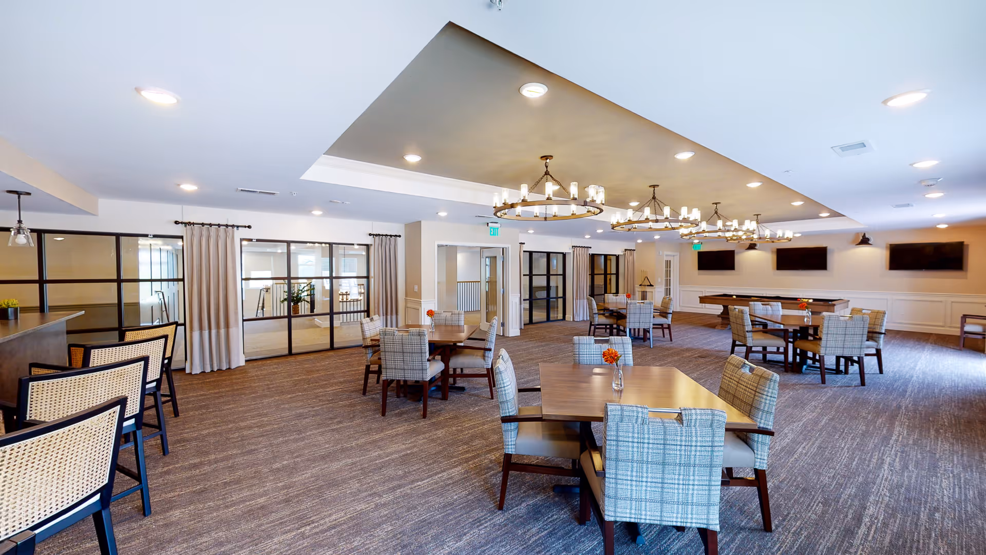 A spacious dining room featuring several tables with chairs, decorative lighting, and large windows with curtains. The room has a warm atmosphere with a neutral color palette and floral centerpieces on the tables.
