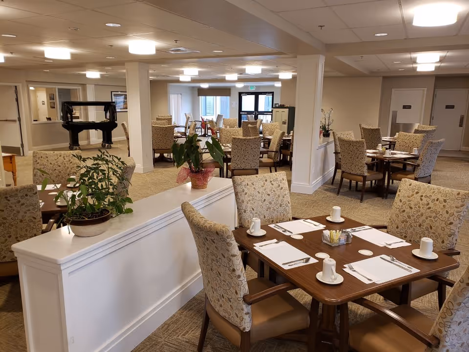 A spacious dining room in a senior living facility with multiple tables set for four people each, featuring patterned cushioned chairs, white placemats, cups, and silverware. The room has beige walls, carpeted floors, ceiling lights, and some potted plants on a white divider. A piano is visible in the background near the entrance doors.