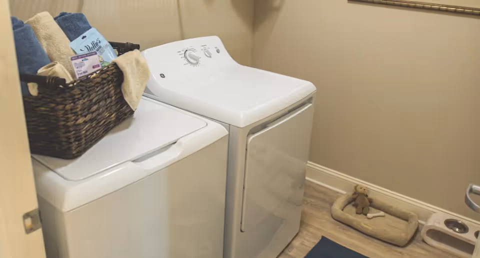 Laundry room with a white washing machine and dryer side by side. A woven basket on top of the washing machine contains folded towels and laundry supplies. On the floor, there is a small pet bed with a stuffed toy and a pet food and water dish nearby. The walls are beige and the floor is wood.