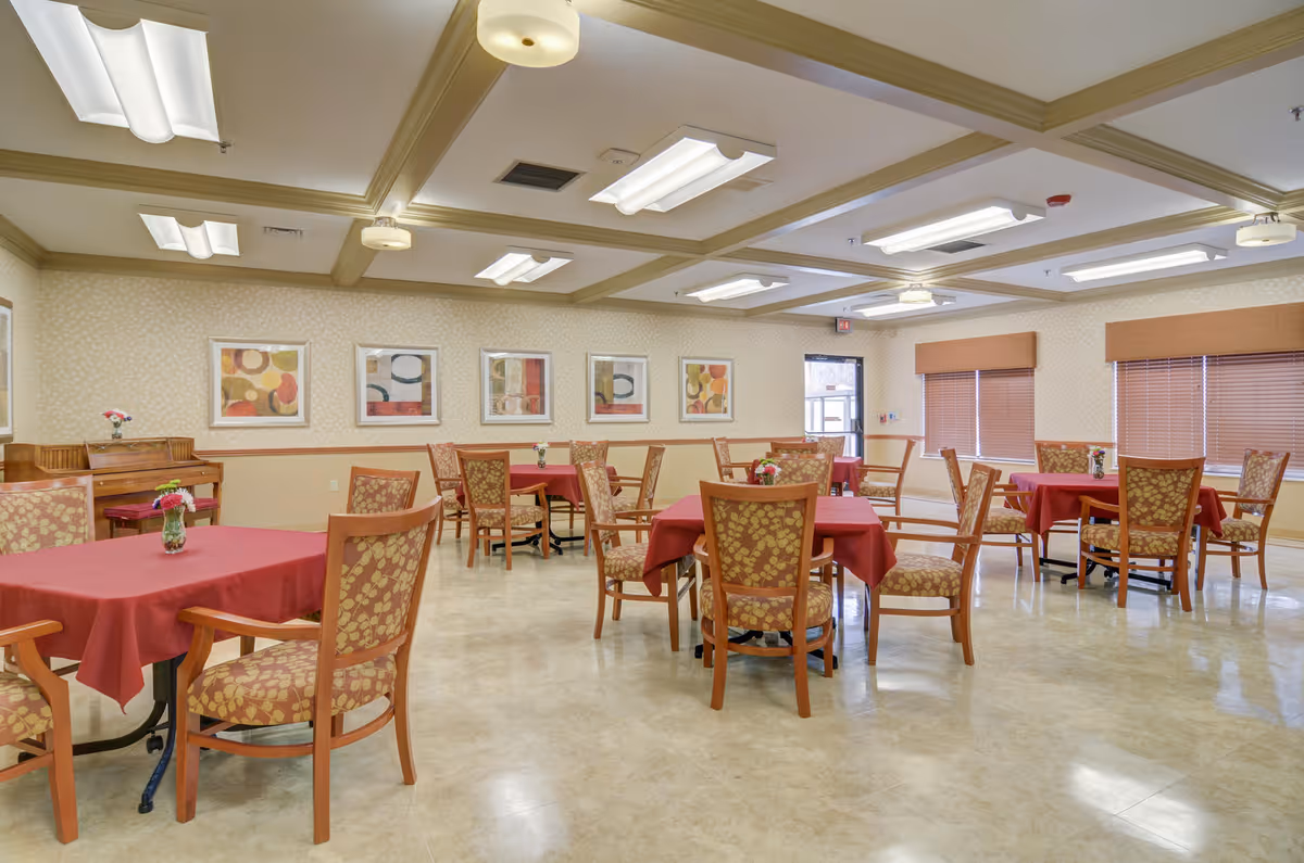 A spacious dining room with multiple square tables covered with red tablecloths, each surrounded by wooden chairs with patterned cushions. The room has beige walls adorned with abstract artwork, large windows with brown blinds, and ceiling lights providing bright illumination. A piano is visible in the corner with a small flower vase on top.