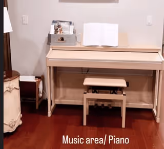 A light-colored upright piano with a matching bench in a room with wooden flooring. On top of the piano, there is an open music book and a small organizer holding additional music sheets. To the left of the piano, there is a small table with a lamp and a magazine rack.