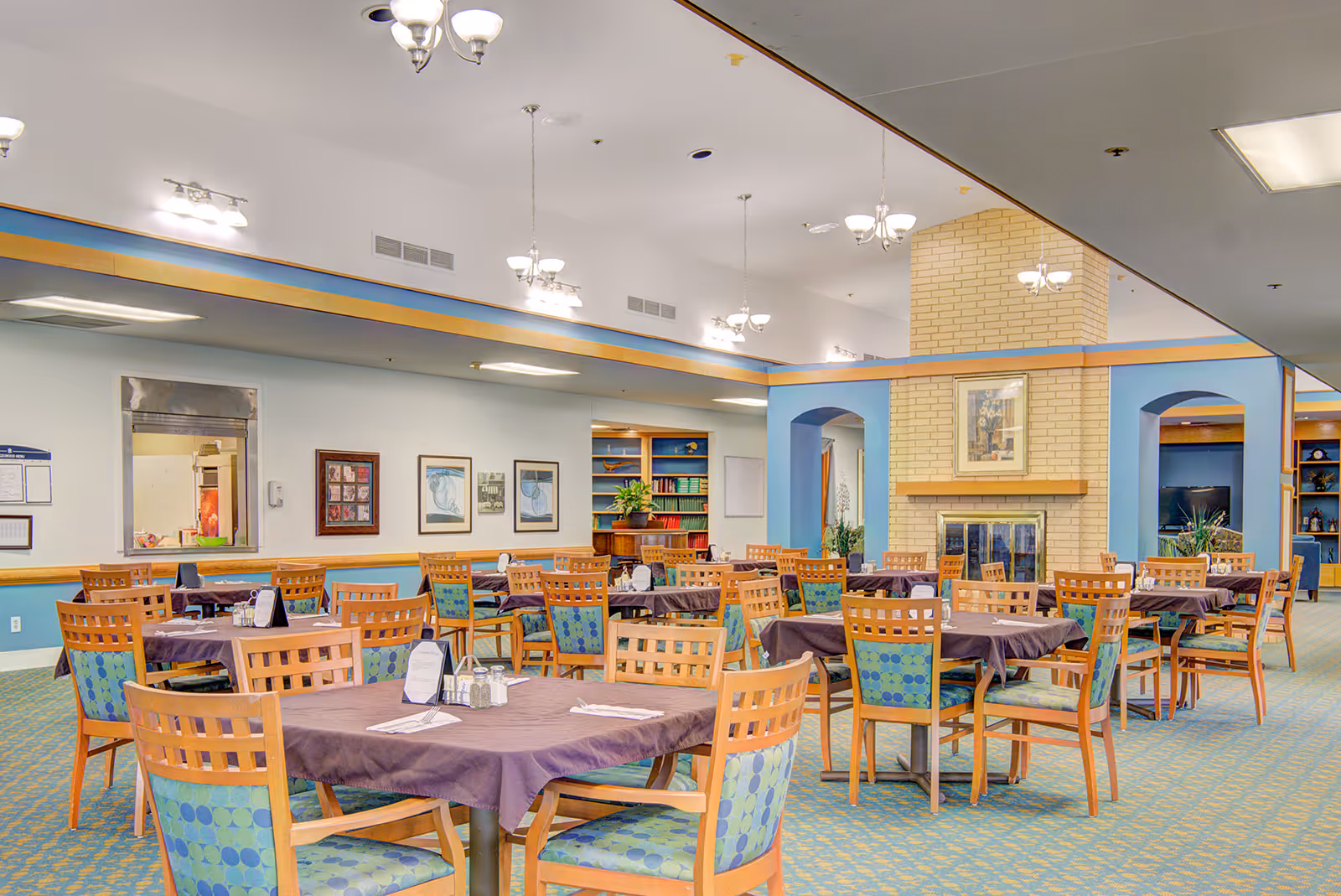 A spacious dining room with multiple wooden tables covered with purple tablecloths and surrounded by wooden chairs with blue and green patterned cushions. The room features a beige brick fireplace with a framed picture above it, blue walls with archways, and several light fixtures hanging from the ceiling. There are framed artworks on the walls and a serving window to the kitchen area.