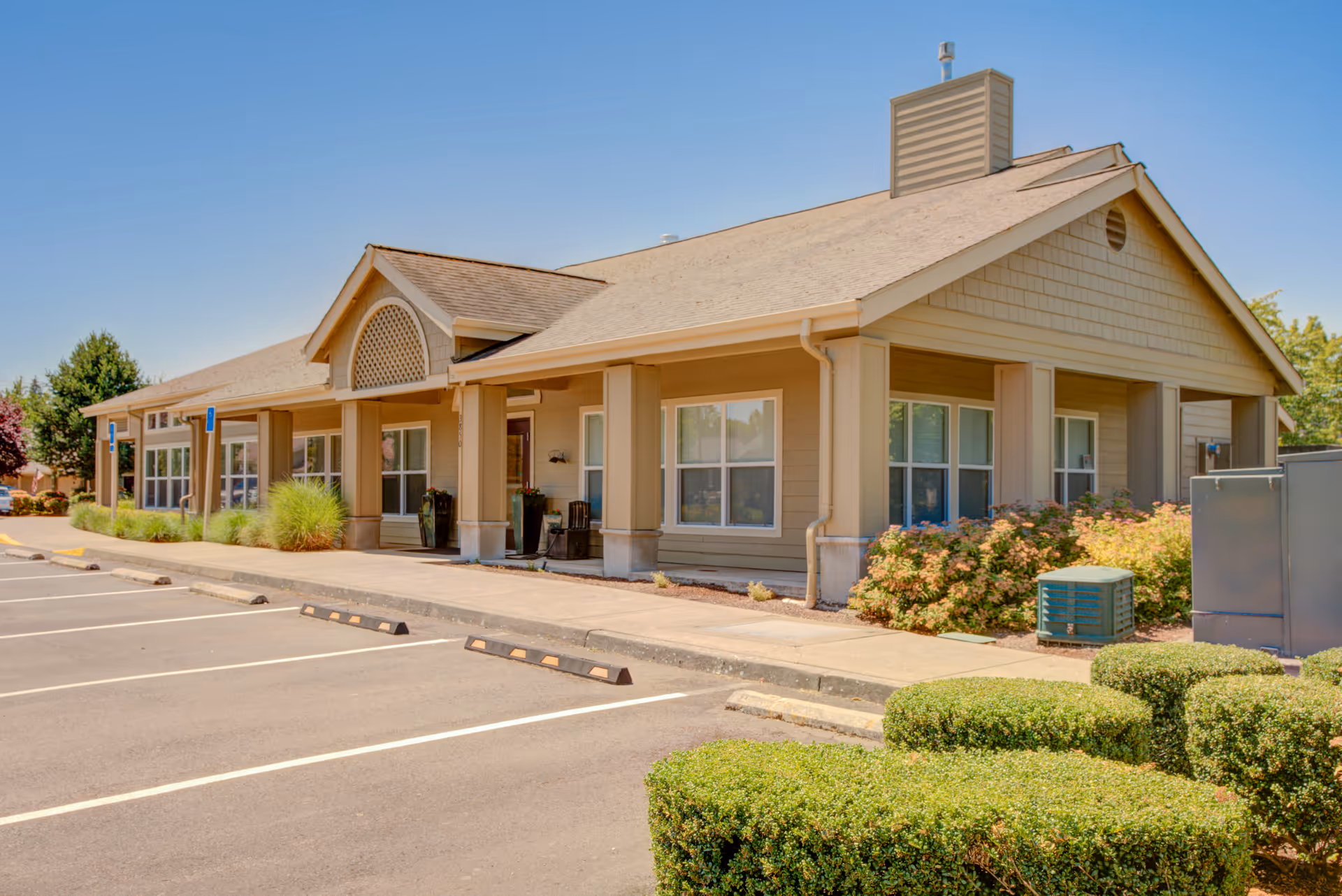 Exterior view of a single-story senior living facility building with beige siding, multiple windows, and a covered entrance. The building is surrounded by neatly trimmed bushes and landscaping, with an empty parking lot in the foreground under a clear blue sky.
