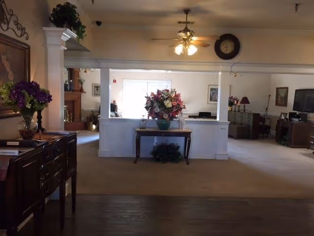 Carpeted common-area interior with a central table holding a large floral arrangement, flanked by white columns and seating areas.