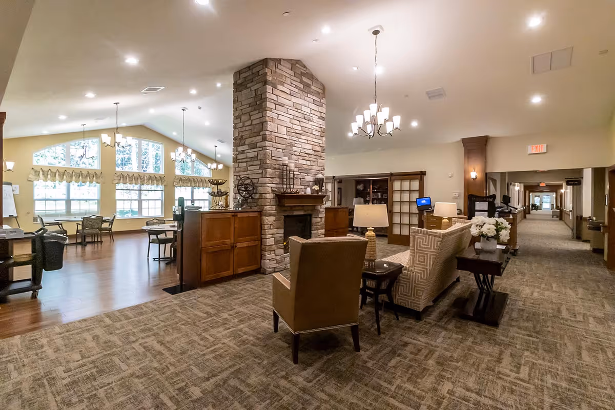 Spacious interior of an assisted living facility featuring a central stone fireplace with wooden cabinets on either side. The room has a seating area with a patterned sofa, an armchair, and a side table with a lamp and flowers. Large windows with valances allow natural light into the dining area with tables and chairs. The ceiling is vaulted with multiple chandeliers and recessed lighting. A hallway extends to the right with exit signs visible.