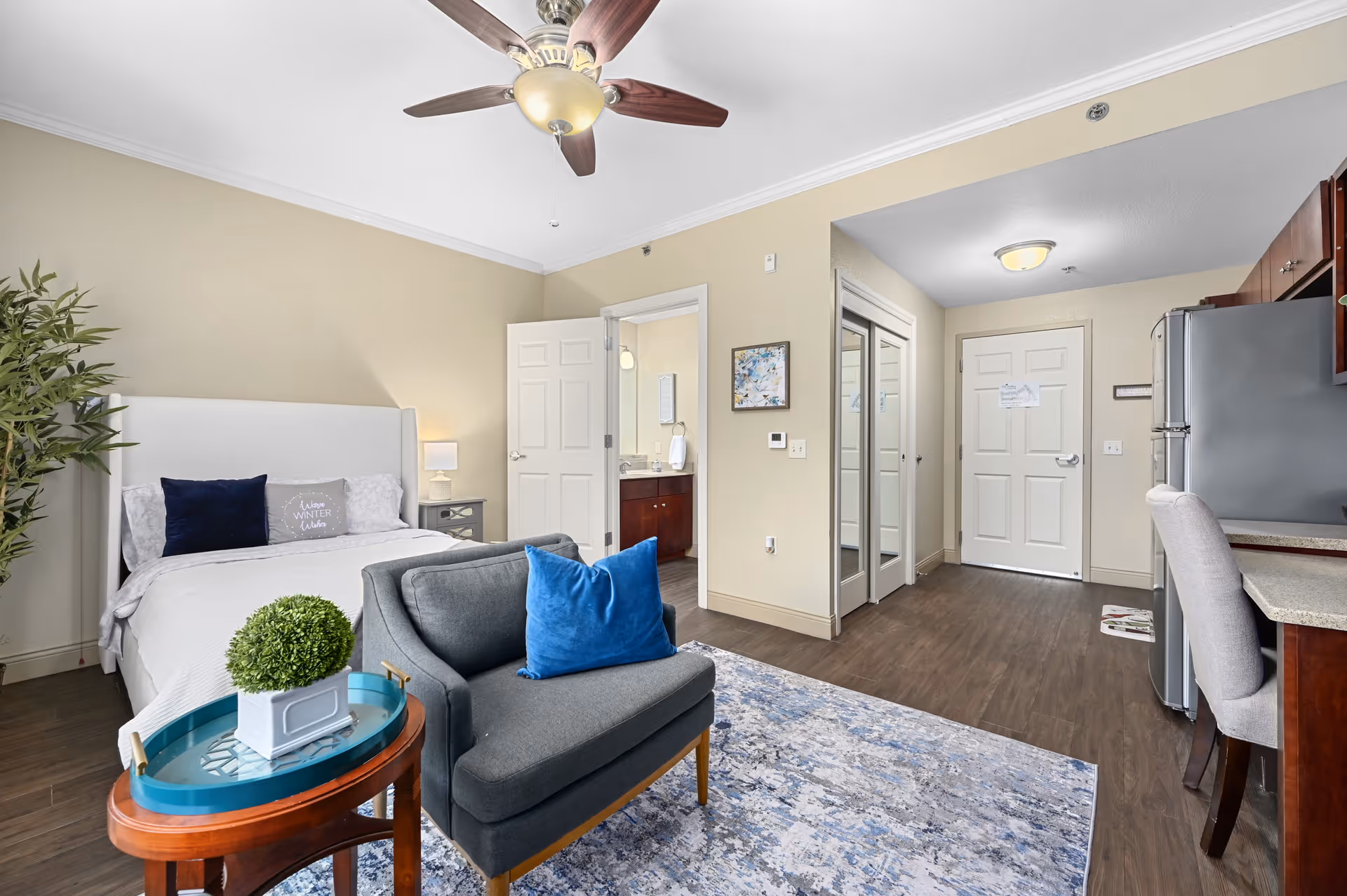 A bright and modern studio apartment featuring a bed with white bedding and decorative pillows, a gray armchair with a blue pillow, a small round wooden side table with a green plant, a ceiling fan with lights, a kitchen area with a refrigerator and countertop, and an open doorway leading to a bathroom. The room has wood flooring, beige walls, and a blue patterned rug.