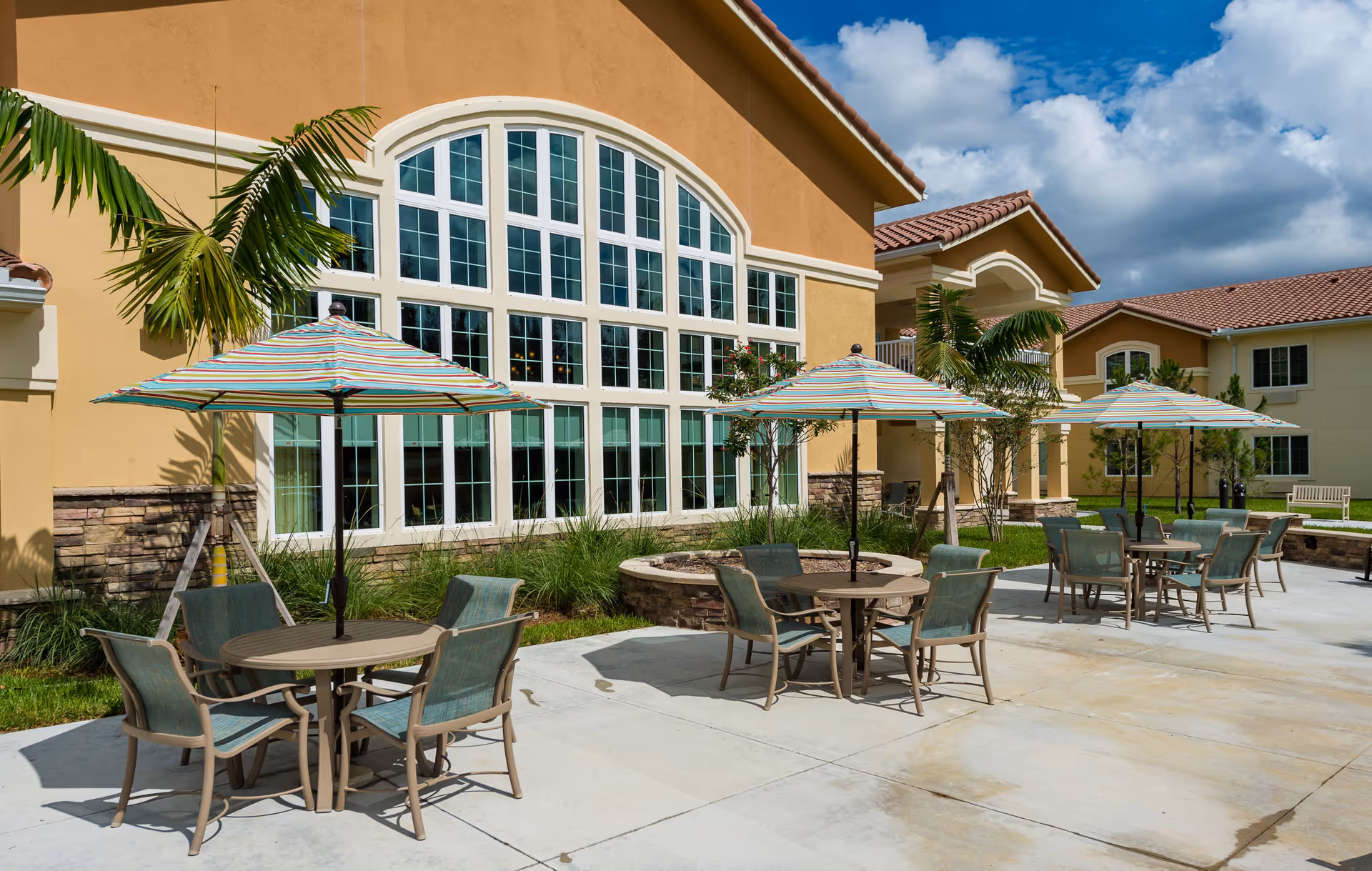 Outdoor patio area at a senior living facility with multiple round tables, each shaded by a striped umbrella. The patio is adjacent to a building with large arched windows and surrounded by palm trees and landscaping under a partly cloudy sky.