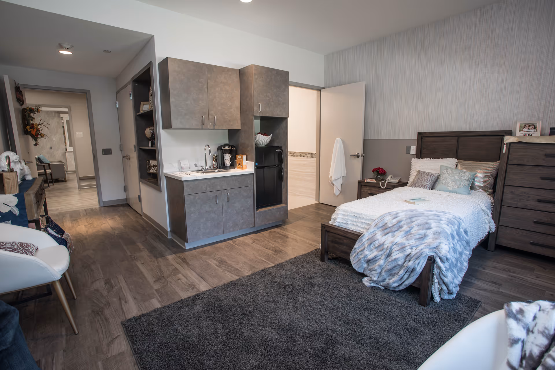 A cozy senior living bedroom featuring a single bed with white and blue bedding, a wooden headboard, and matching dresser. The room includes a small kitchenette with cabinets, a sink, a coffee maker, and a mini refrigerator. There is a gray rug on the wooden floor, a white chair with a pillow, and an open door leading to a bathroom.