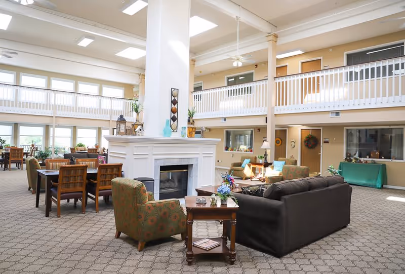 Spacious senior living facility common area with a central white fireplace, surrounded by comfortable seating including a dark sofa and patterned armchairs. There are wooden tables and chairs arranged for dining or socializing, large windows letting in natural light, and a second-floor balcony with white railings overlooking the area.