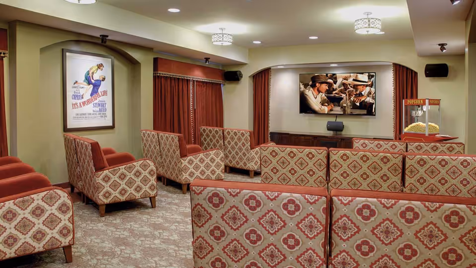 A cozy movie theater room with patterned upholstered chairs arranged in rows facing a large flat-screen TV mounted on the wall. The room has warm lighting, red curtains, a popcorn machine filled with popcorn on the right side, and a vintage movie poster on the left wall.