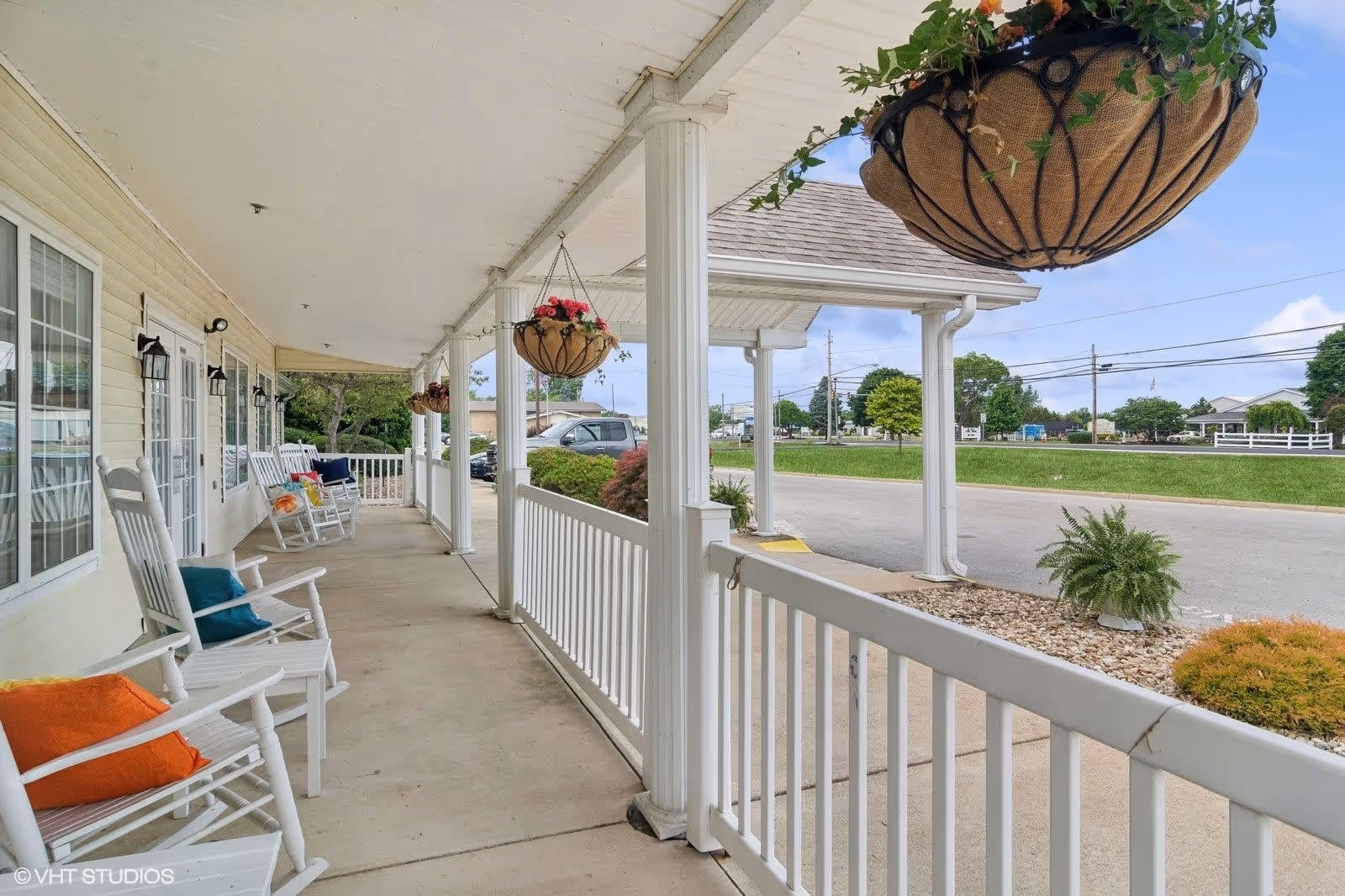 Covered porch area with white rocking chairs adorned with colorful cushions, hanging flower baskets, white railing, and a view of a parking lot and greenery in the background under a partly cloudy sky.