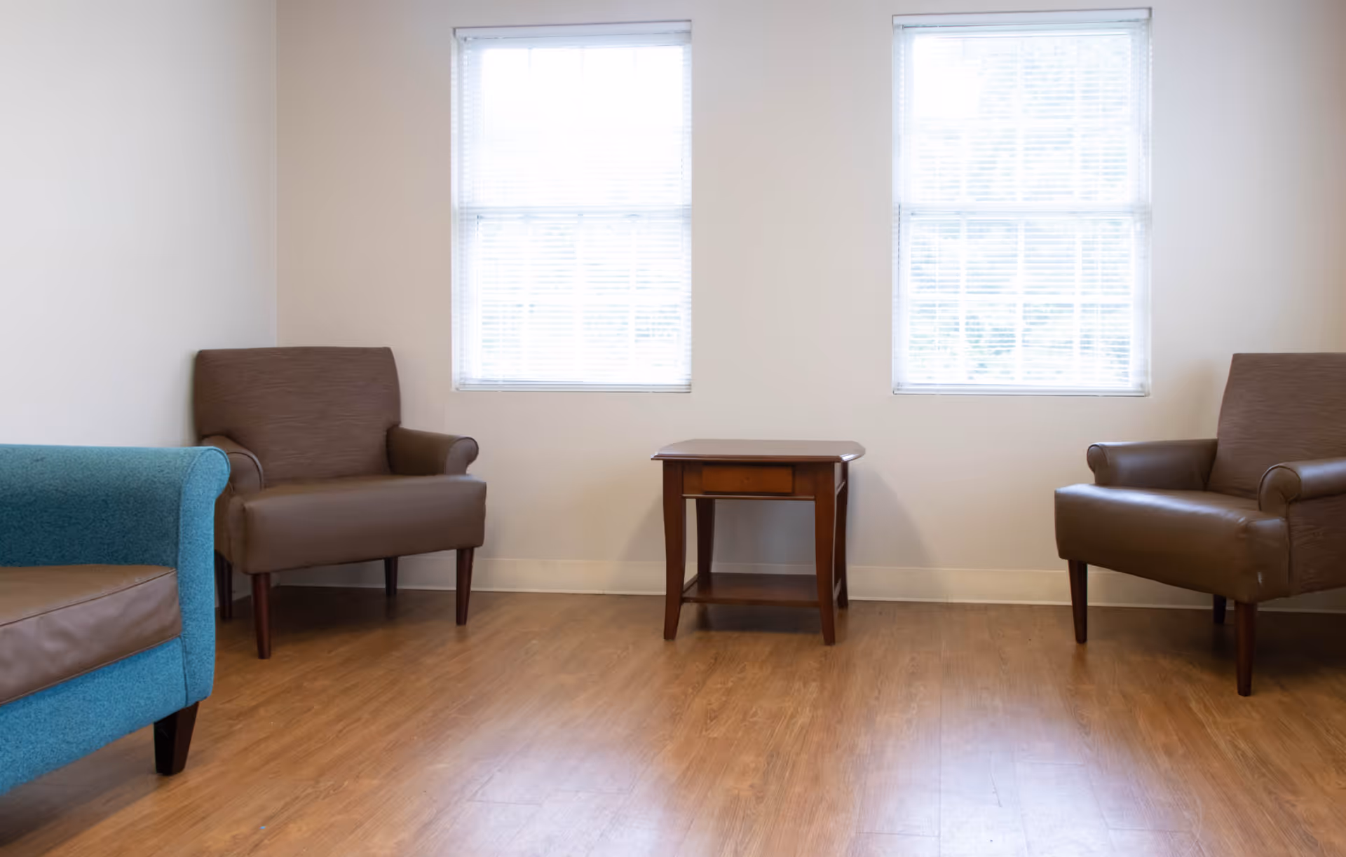 A simple sitting area with two brown armchairs and one blue armchair arranged around a small wooden side table, with two windows with white blinds on the wall behind them.
