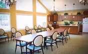 A bright dining room with large windows letting in natural light. The room features a long rectangular table surrounded by cushioned chairs with wooden frames. In the background, there is a kitchen area with wooden cabinets, a refrigerator, and a countertop with plants and decorative items. The walls are painted yellow, and chandeliers hang from the ceiling.