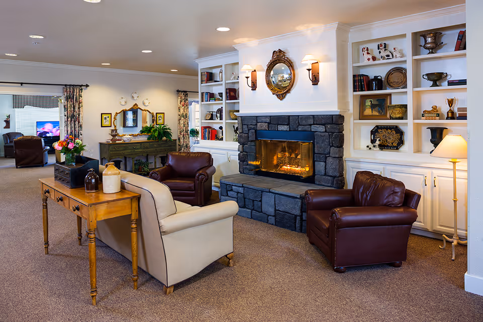 A cozy living room area in a memory care community featuring a stone fireplace with a fire burning, flanked by built-in white shelves filled with books and decorative items. The room has two brown leather armchairs and a beige sofa arranged around the fireplace. A wooden console table with decorative vases is placed behind the sofa. The walls are adorned with a round mirror above the fireplace and framed pictures. In the background, there is another seating area with a television and floral curtains on the windows.