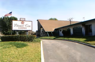 Exterior view of a single-story building with a sloped roof and a driveway leading up to it. There is a sign on the lawn that reads 'Pleasant Care CORONA' with an American flag flying on a pole nearby. The sky is clear and the surrounding area has green grass and some bushes.