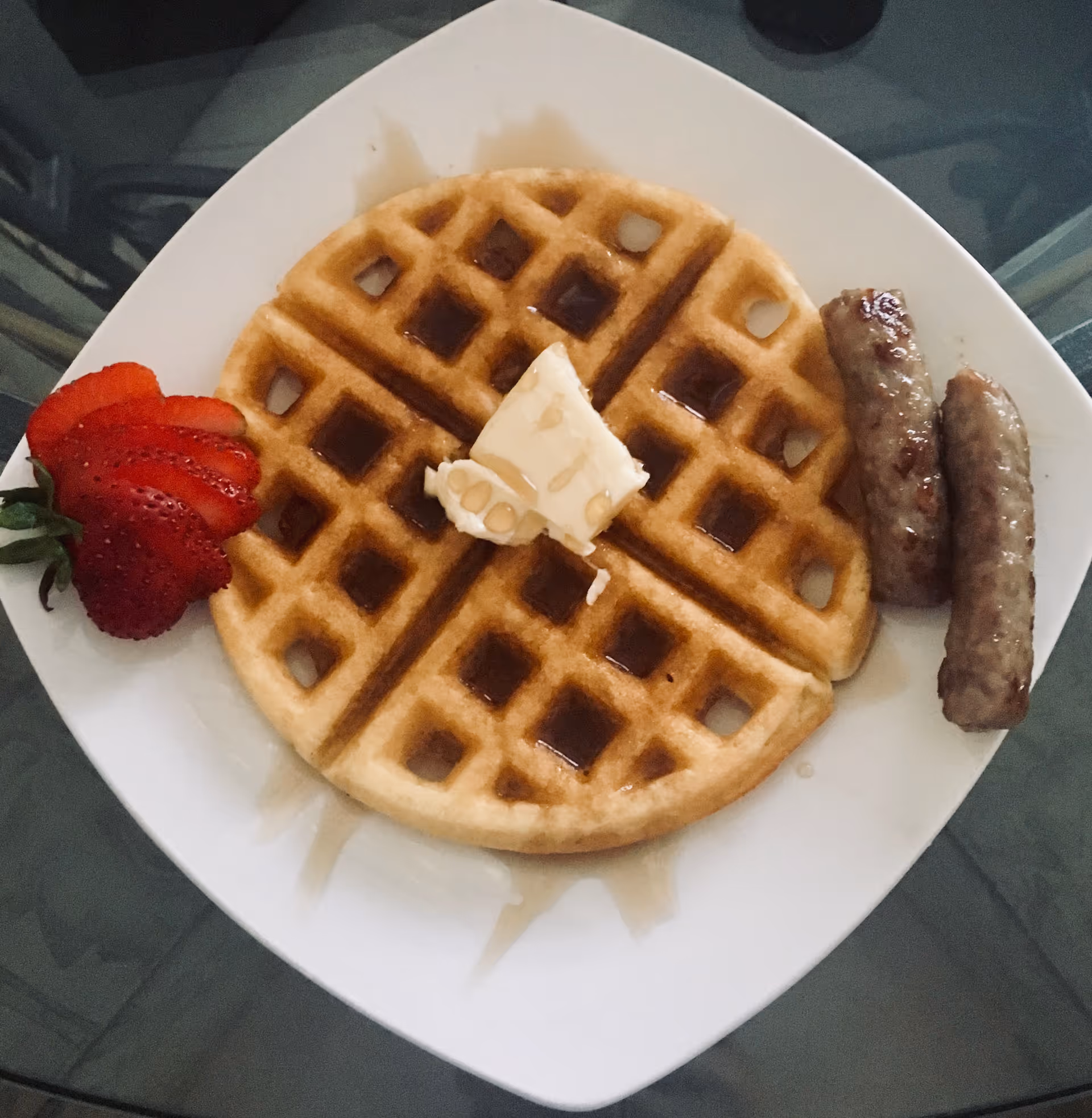 A white square plate with a round waffle topped with a pat of butter and syrup, two sausage links on the side, and a sliced strawberry garnish.