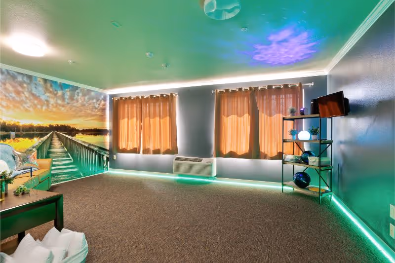 A cozy living room with gray walls and carpeted floor, featuring three windows with brown curtains. There is a large mural of a wooden pier extending into a lake at sunset on one wall. The room has green LED strip lighting along the baseboards and ceiling edges. A small black metal shelving unit holds decorative items and a small TV mounted on the wall. A wooden coffee table with a plant and pillows is partially visible.