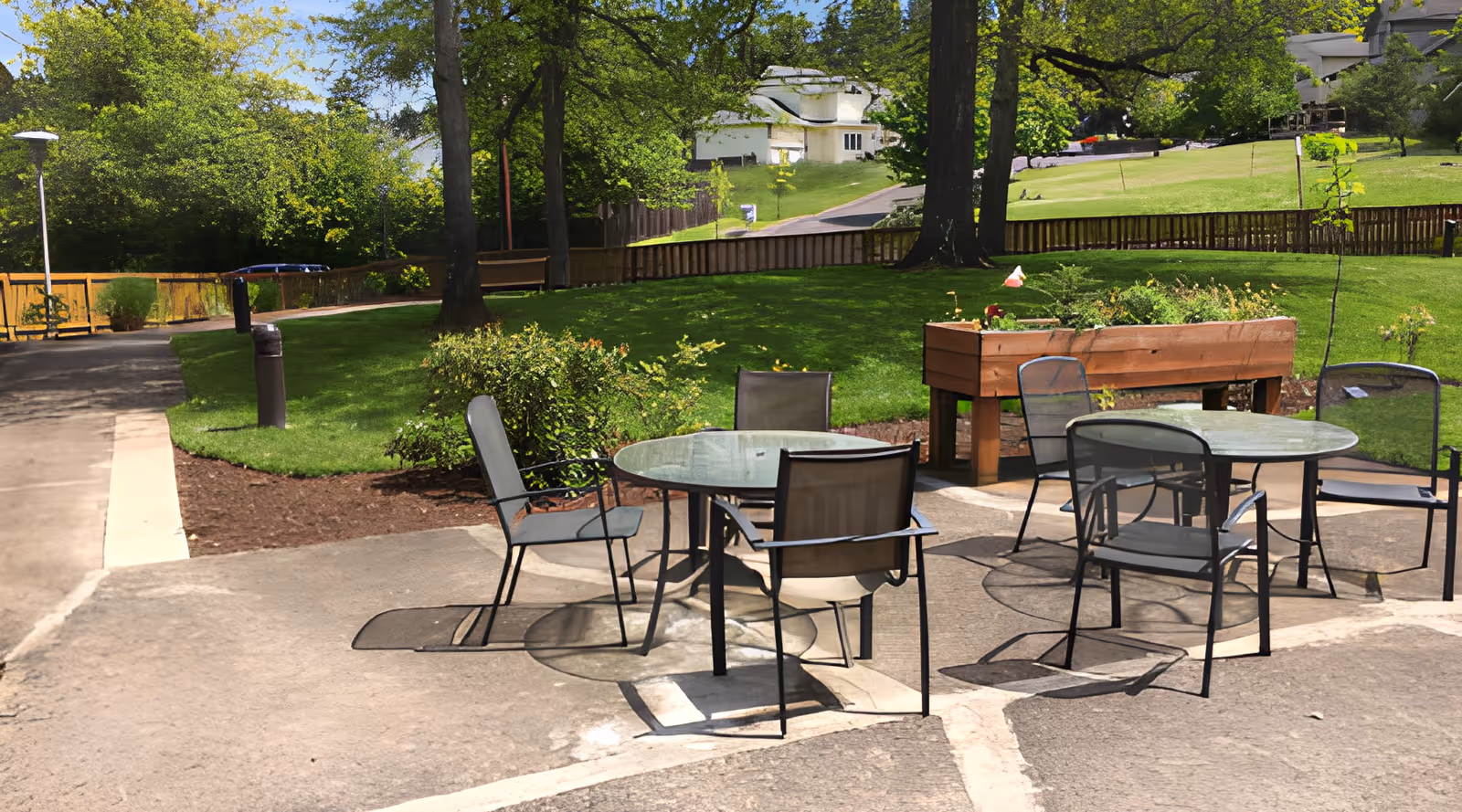 Outdoor patio area with two round glass tables and several metal chairs on a concrete surface, surrounded by green grass, trees, and a wooden raised garden bed, with a pathway and residential houses in the background.