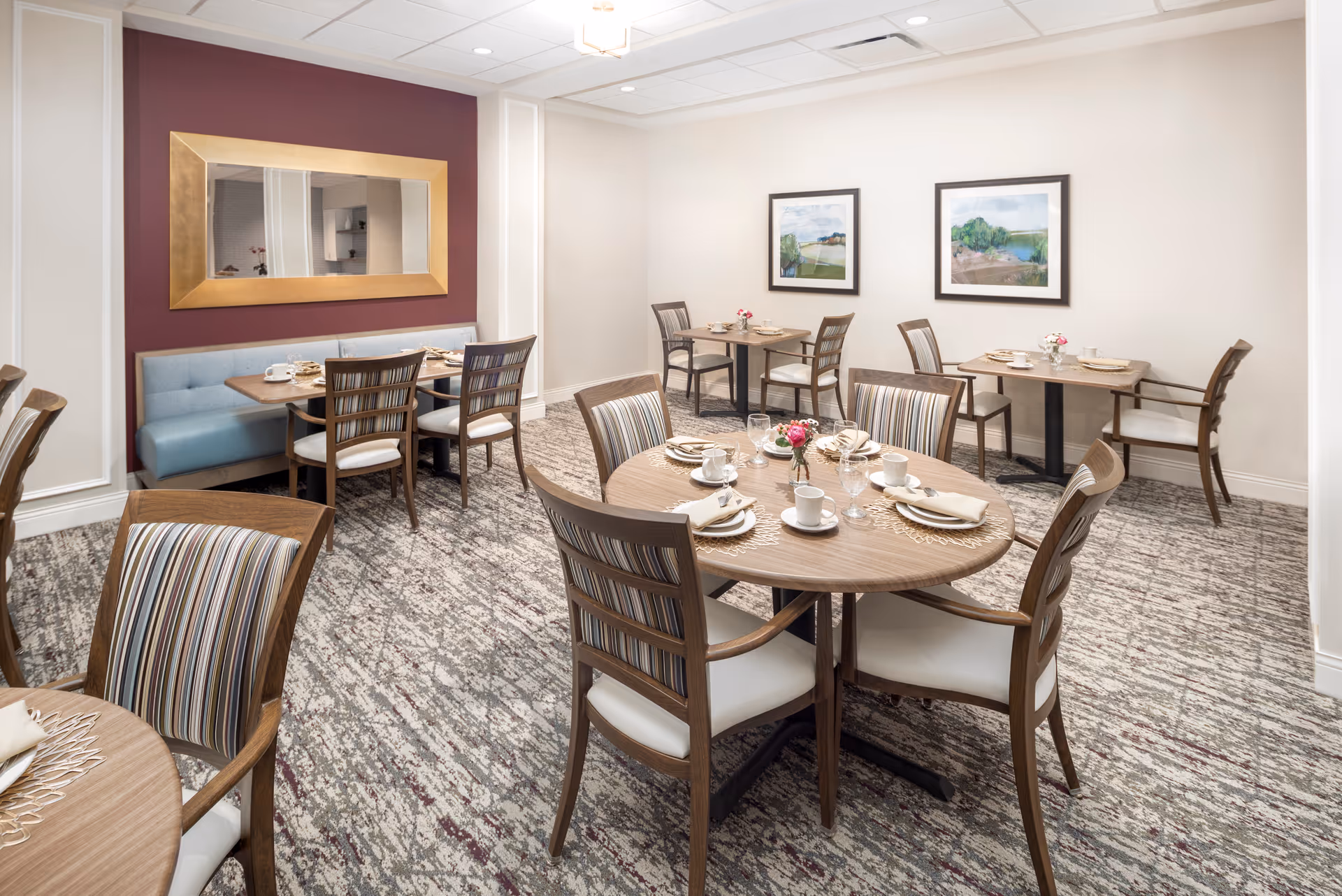 A dining room in a senior living facility with several wooden tables and chairs arranged neatly. The tables are set with white cups, plates, napkins, and small flower vases. The room has a patterned carpet, light-colored walls, two framed landscape paintings, and a large gold-framed mirror on a maroon accent wall.