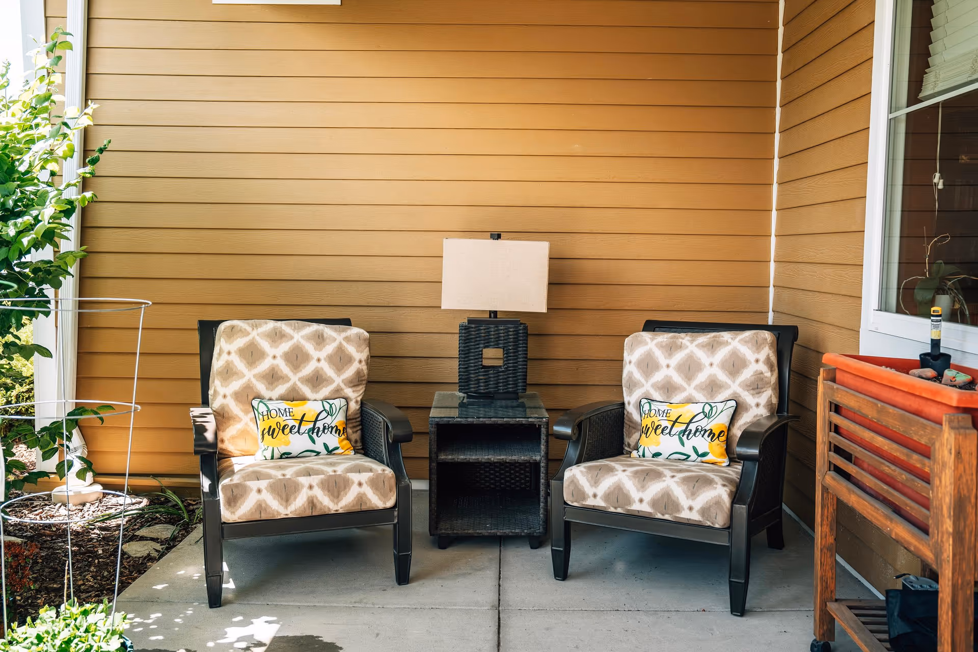 Two cushioned armchairs with patterned beige upholstery and 'Home Sweet Home' pillows are placed on a porch with a small black wicker table and lamp between them. The porch has tan horizontal siding, a window with blinds, and some plants nearby.