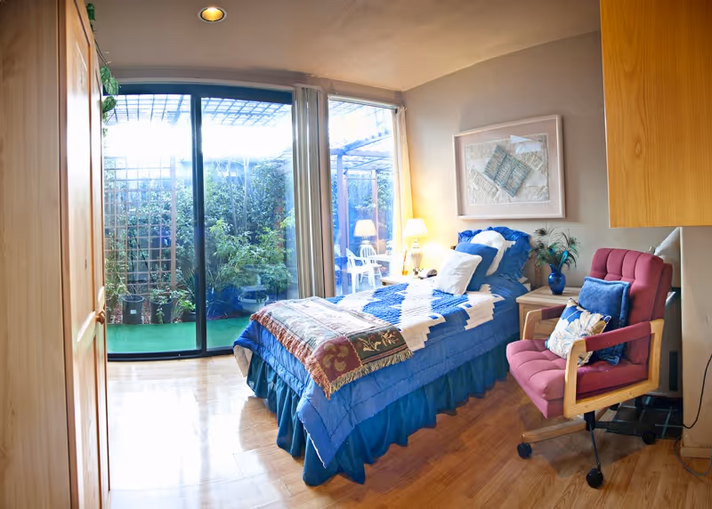 A cozy bedroom with a single bed covered in blue bedding and a decorative throw. Next to the bed is a wooden nightstand with a lamp and a vase with flowers. A red cushioned chair with pillows is positioned near the bed. The room has wooden flooring and a large sliding glass door that opens to a small outdoor garden area with plants and a white chair. Light beige walls and a framed artwork hang above the bed.
