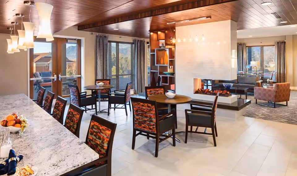 A bright and spacious common area in a senior living facility featuring a marble countertop with chairs, round tables with patterned chairs, a modern double-sided fireplace, large windows with curtains, and a cozy seating area with sofas and armchairs.