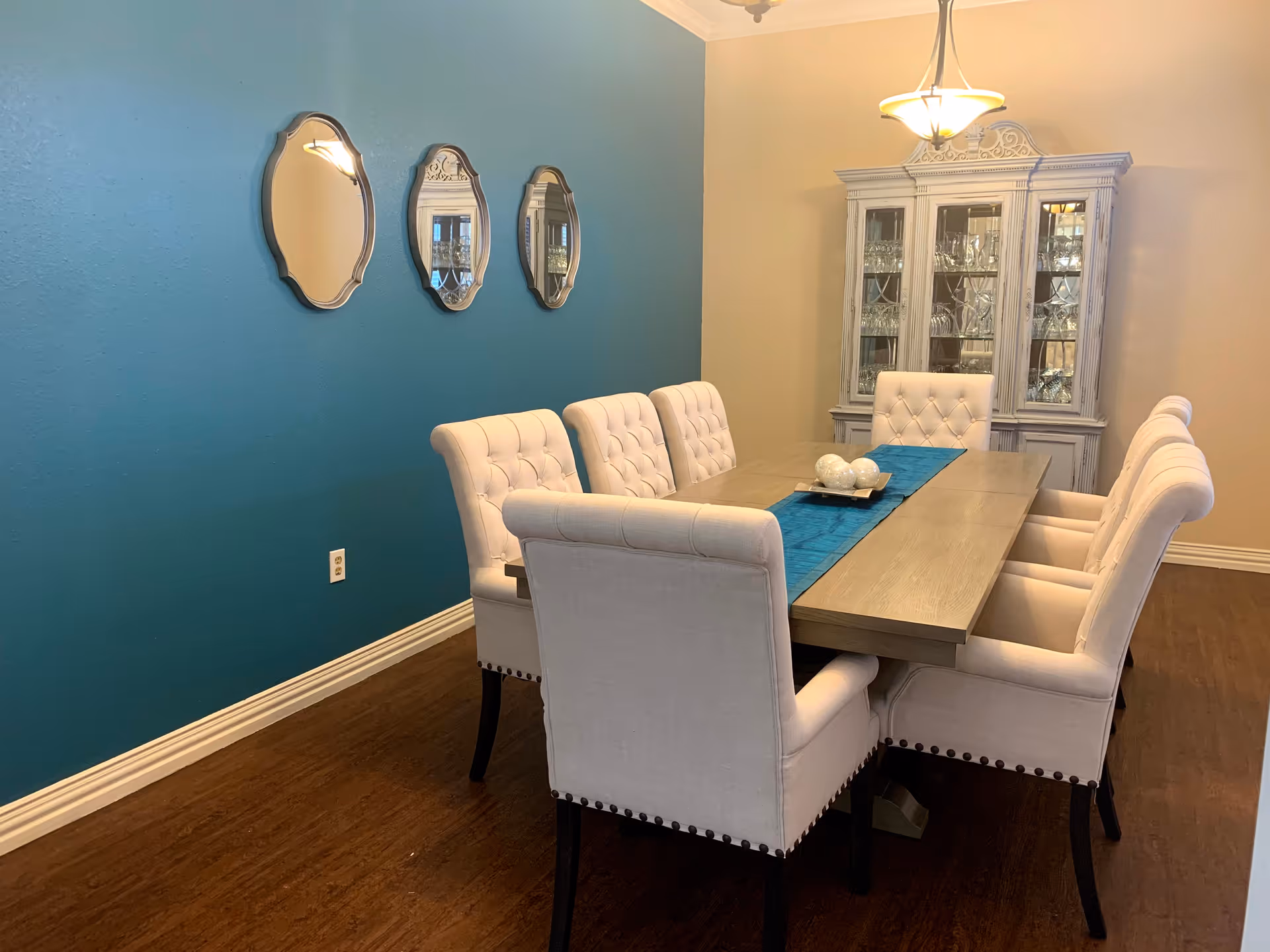 Dining room with a long wooden table surrounded by upholstered chairs, a teal accent wall with three mirrors, and a glass-front china cabinet.