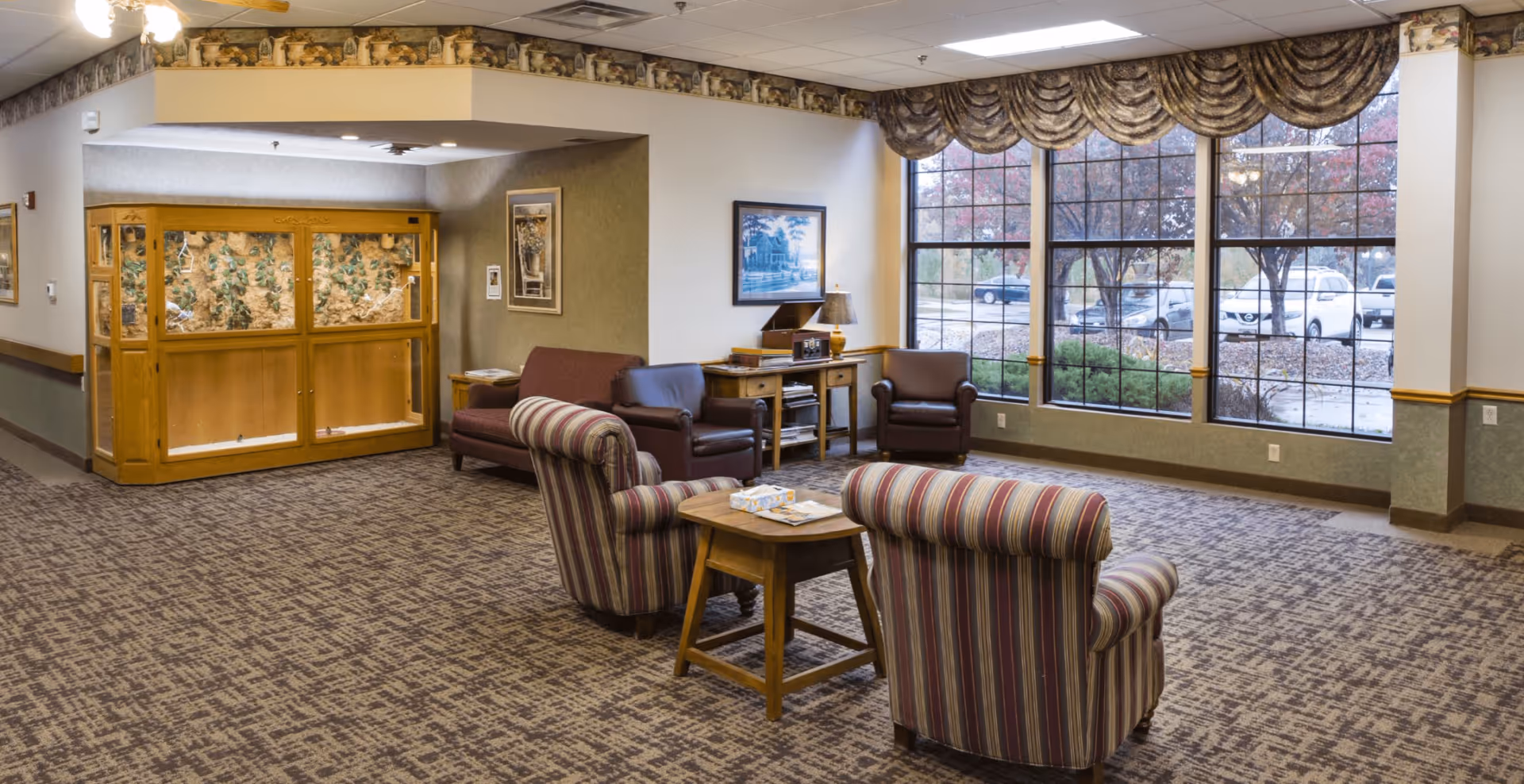 A cozy seating area in a senior living facility with two striped armchairs and a wooden side table in the foreground, two leather armchairs and a loveseat against the wall, a wooden cabinet with a lamp and framed picture, large windows with decorative valances showing a parking lot and trees outside, and a large wooden display case with plants on the left side.