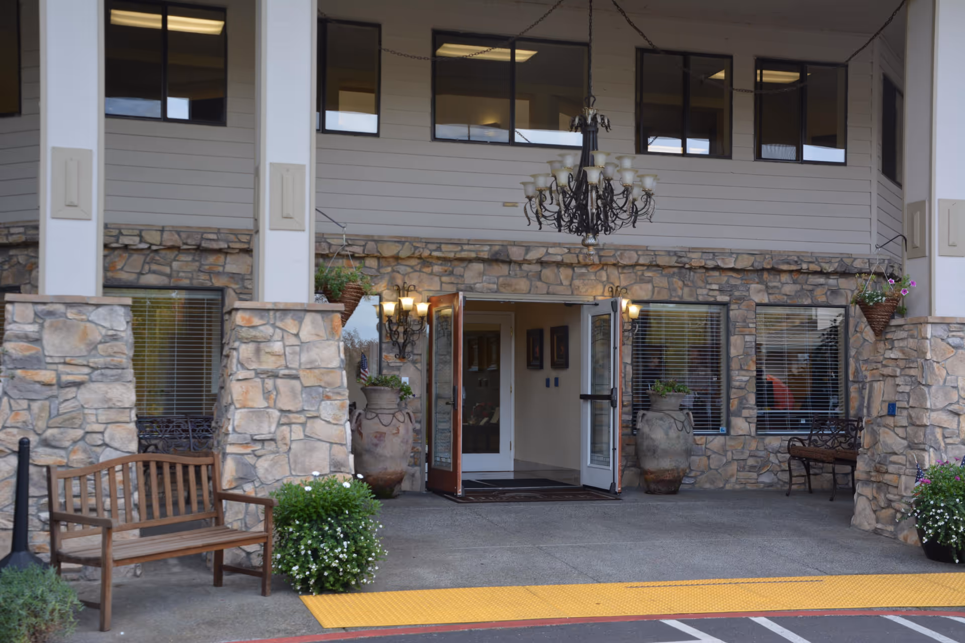 Entrance of a building with stone walls and white pillars, featuring double glass doors that are open. There are two large decorative urns with plants on either side of the entrance, a wooden bench to the left, and a metal bench to the right. A large chandelier hangs from the ceiling above the entrance, and there are wall-mounted lights on both sides of the doors. The ground has a yellow tactile paving strip near the entrance.