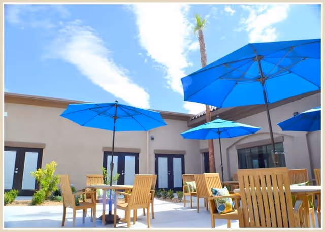 Sunny outdoor courtyard with wooden tables and chairs under bright blue umbrellas beside a low building.