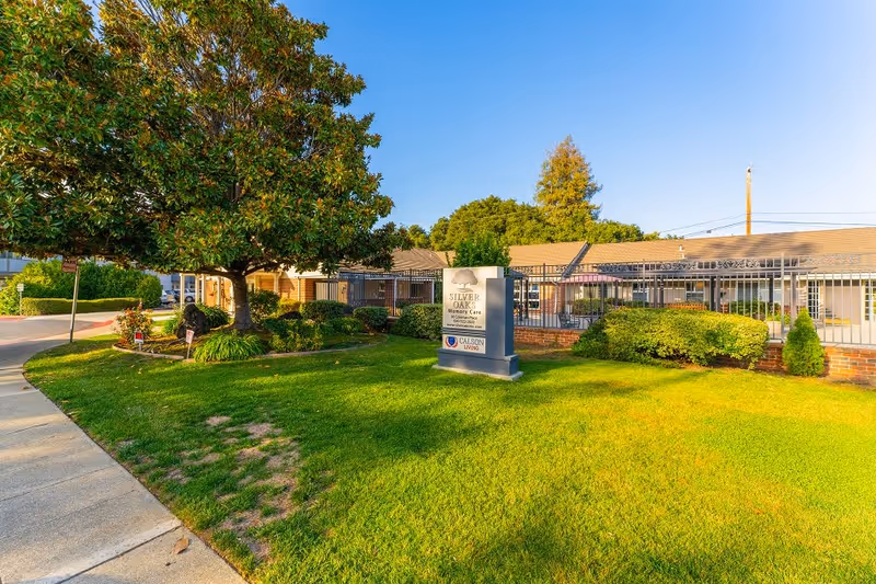Exterior view of Silver Oaks Memory Care facility with a large green lawn, a tree, bushes, and a sign displaying the facility's name and logo. The building is single-story with a fenced area and a clear blue sky above.