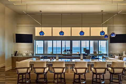 Modern dining area with a large rectangular table surrounded by cushioned chairs, pendant lights with blue glass shades hanging above, and large windows offering a view of the ocean.