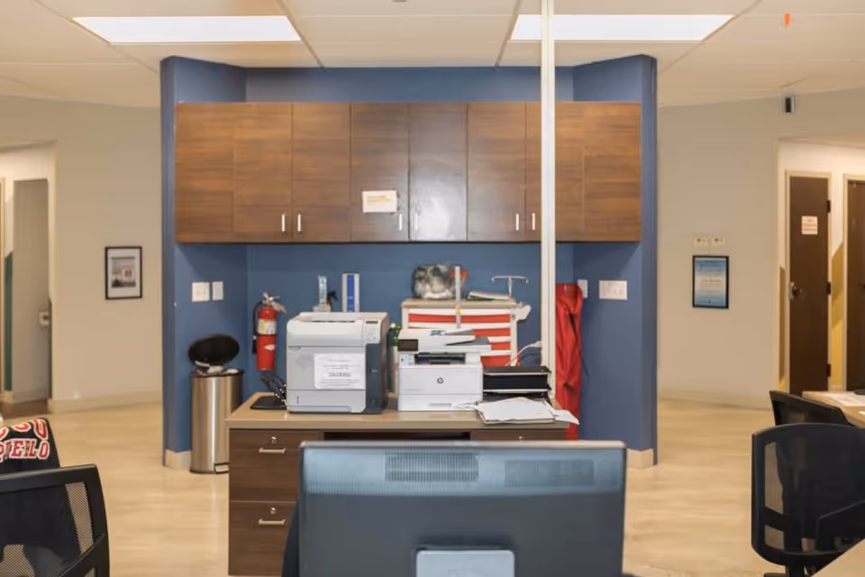 A clinical reception/workstation area with printers, wood cabinets, office chairs and a blue accent wall.