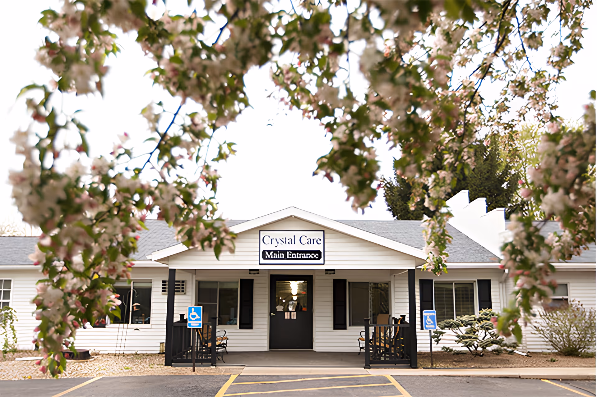 Front exterior view of Crystal Care Center Of Ashland building with a covered entrance, two benches on either side, handicap parking signs, and flowering tree branches framing the top of the image.