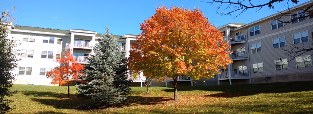Front of a multi-story senior living building with autumn trees and a grassy lawn under a clear blue sky.