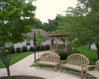 Outdoor garden area with wooden benches and a gazebo surrounded by trees and shrubs, with a building visible in the background.